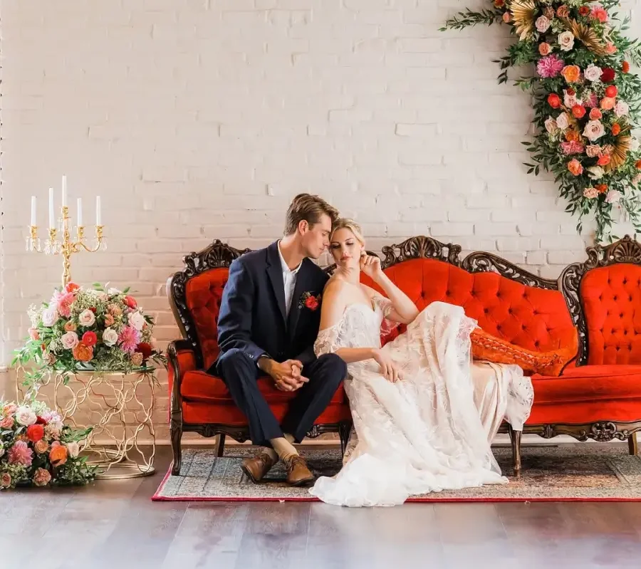 Wedding couple relax on a long vintage red velvet sofa, surrounded by flowers and elegant side pieces