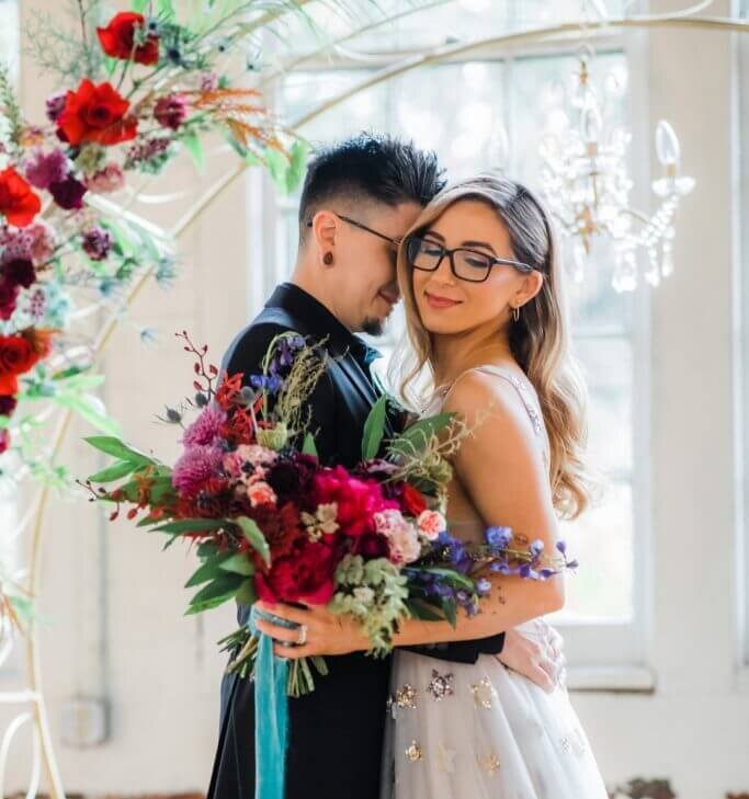 A wedding couple stand beneath a white metal arbor trimmed with jewel toned flowers